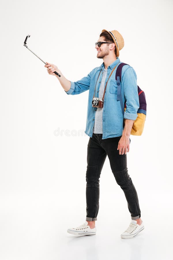 Smiling attractive young tourist with backpack walking and using mobile phone on selphie stick over white background. Smiling attractive young tourist with backpack walking and using mobile phone on selphie stick over white background