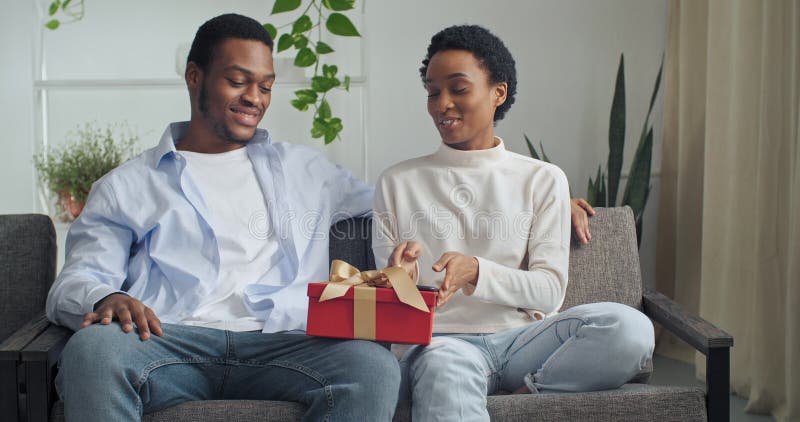 Smiling two people afro american couple short hair woman and black man sitting together at home sofa holding red gift