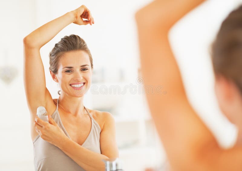 Smiling young woman applying roller deodorant on underarm in bathroom