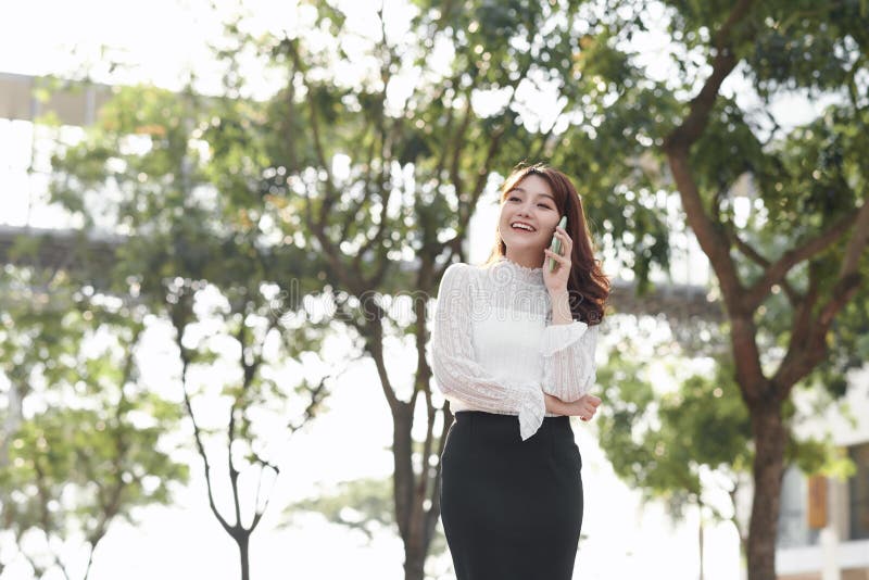 Smiling woman calling on phone outdoors. Pretty young lady standing with city view in background. City walk concept. Side view royalty free stock photo Smiling woman calling on phone outdoors. Pretty young lady standing with city view in background. City walk concept. Side view royalty free stock photo