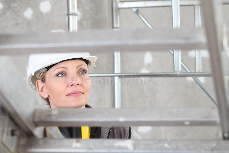 Smiling woman construction worker builder on ladder wearing white helmet and hearing protection headphones on interior site stock photo