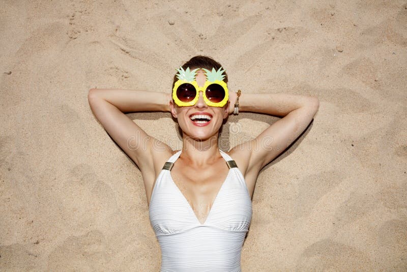 Smiling woman in swimsuit and pineapple glasses laying on sand