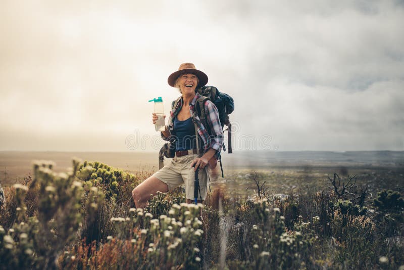 Smiling woman taking a break while hiking in the countryside drinking water. Cheerful senior woman wearing backpack and hat standing on a hill holding a water bottle. Smiling woman taking a break while hiking in the countryside drinking water. Cheerful senior woman wearing backpack and hat standing on a hill holding a water bottle