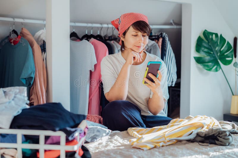 Smiling woman using smartphone to find places where to donate or sale clothes. Decluttering, Sorting clothes and Cleaning Up. Conscious consumer, sustainability. Reuse, second hand, Charity concept
