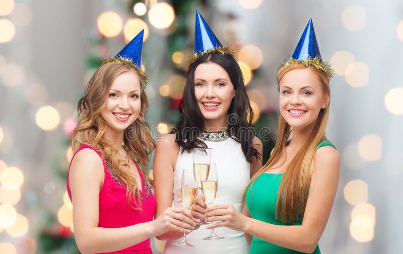 Drinks, holidays, people and celebration concept - smiling women in party hats with glasses of sparkling wine over christmas tree lights background. Drinks, holidays, people and celebration concept - smiling women in party hats with glasses of sparkling wine over christmas tree lights background