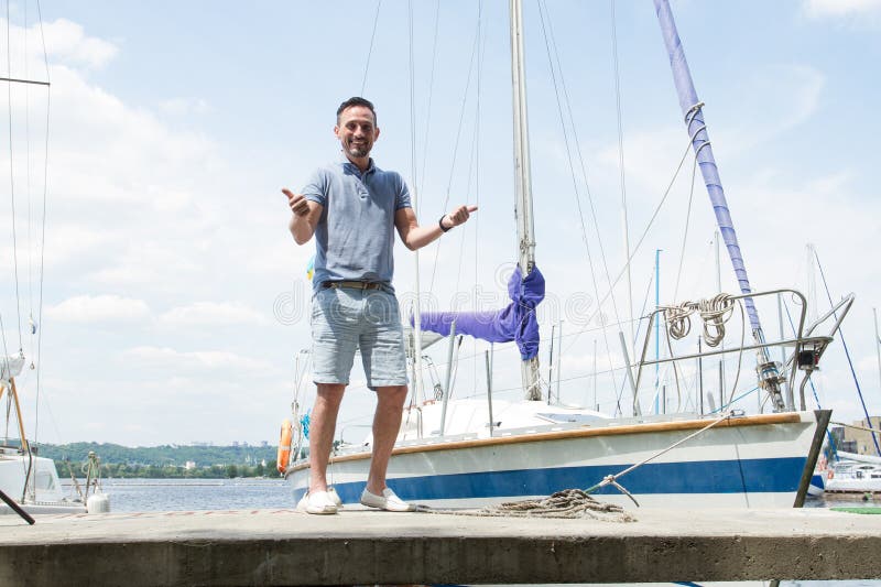 A Smiling Yachtsman portrait on pier with both thumbs up. river and yachts on background. Young successful man sailor on pier
