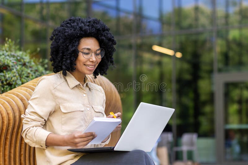 Smiling young African American female student sitting on a bench on a university campus studying online using a laptop. Writes in a notebook and listens to a lecture.