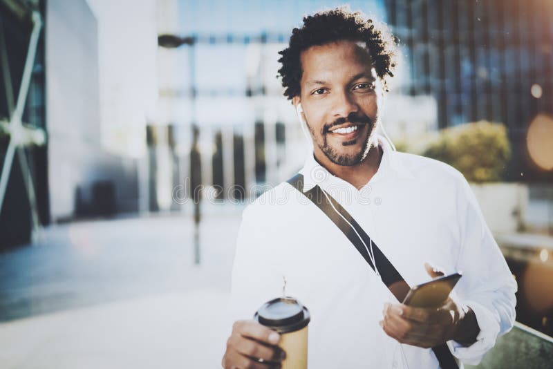 Smiling young African American man in headphone walking at sunny city with take away coffee and enjoying to listen to