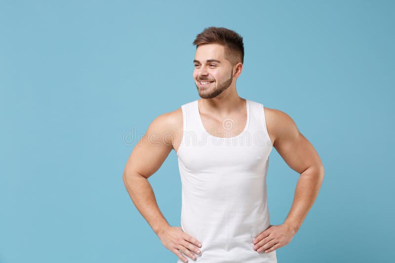 Smiling young bearded guy 20s in white singlet posing isolated on pastel blue background. Sport fitness healthy lifestyle concept. Mock up copy space. Standing with arms akimbo on waist looking aside