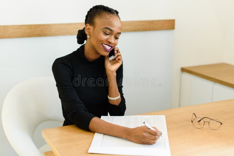 Smiling young black business woman on phone taking notes in office stock photo Smiling young black business woman on phone taking notes in office stock photo
