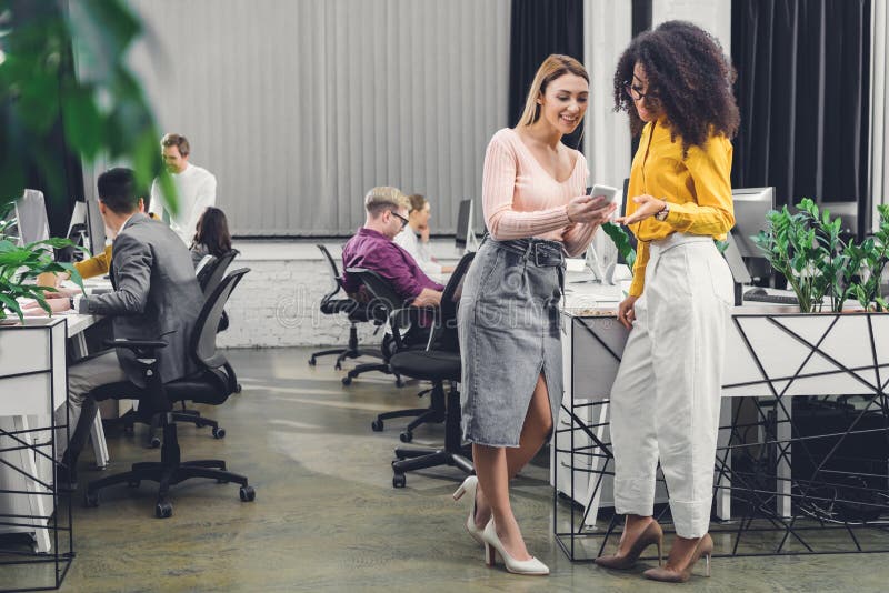 Smiling young businesswomen using smartphone and talking while colleagues working behind stock photo Smiling young businesswomen using smartphone and talking while colleagues working behind stock photo