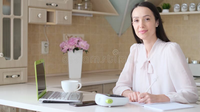 Smiling young freelancer woman working with laptop in kitchen and taking notes. stock video footage