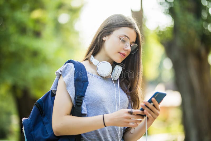 Smiling young girl student with backpack holding mobile phone with earphones, walking at the park royalty free stock photo Smiling young girl student with backpack holding mobile phone with earphones, walking at the park royalty free stock photo