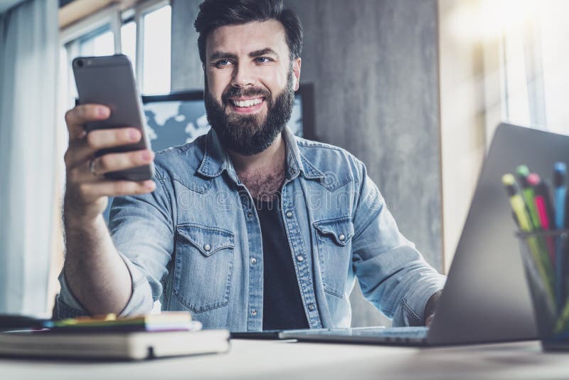 Young man sitting at window and working on laptop. Bearded man sitting at  desktop with smartphone in his hands. Online education. Stock Photo