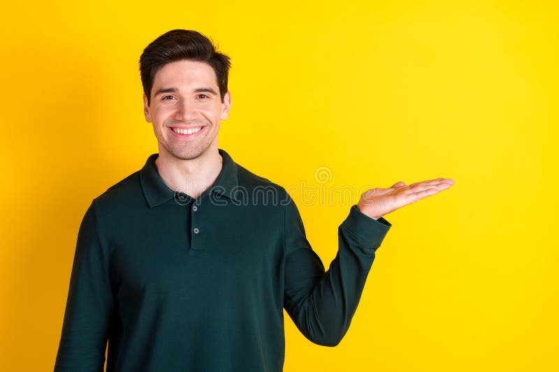 Smiling young man in a green polo shirt posing against a yellow background while gesturing with his hand in a presenting royalty free stock photos