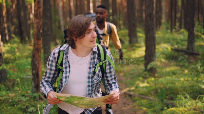 Smiling young man is looking at map and searching for right way in forest while multiethnic group of friends is