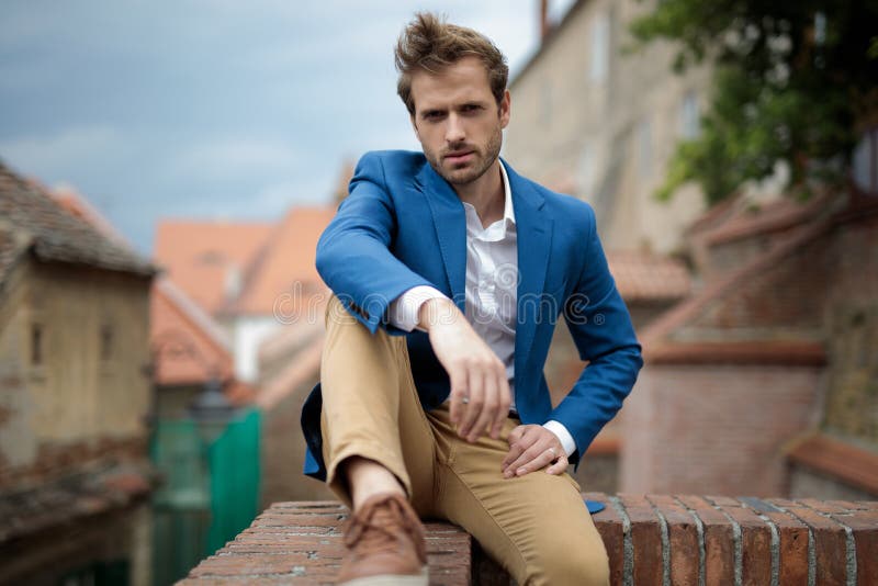 Smiling young sexy man sits of a brick wall with old city behind