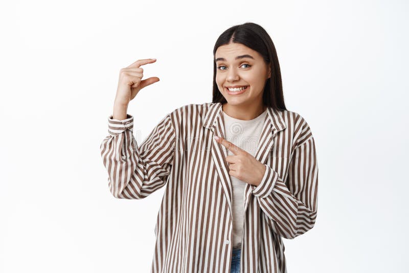 Smiling young woman awkwardly pointing at small size gesture, showing little thing, tiny item in hand, standing against white background.