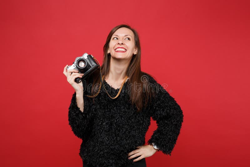 Smiling young woman in black fur sweater looking up, holding retro vintage photo camera isolated on bright red wall stock photography Smiling young woman in black fur sweater looking up, holding retro vintage photo camera isolated on bright red wall stock photography