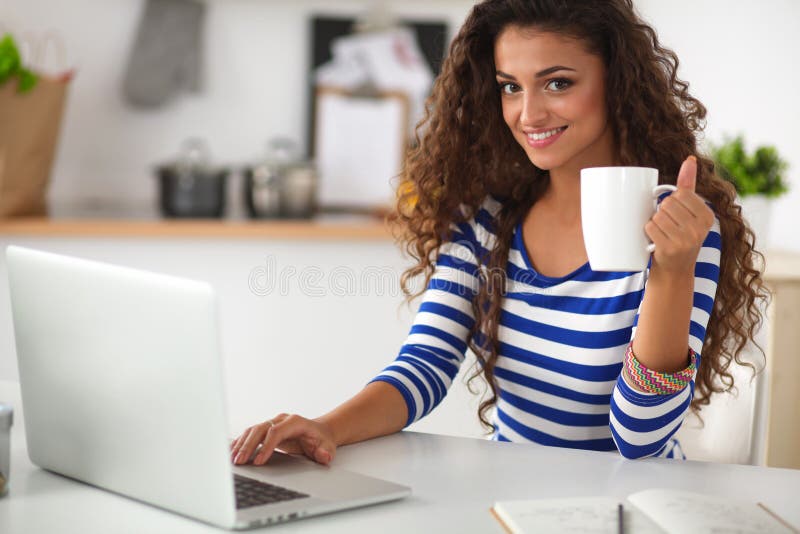 Smiling young woman with coffee cup and laptop in the kitchen