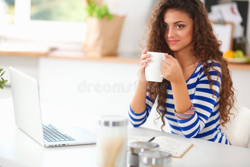 Smiling young woman with coffee cup and laptop in the kitchen at home.