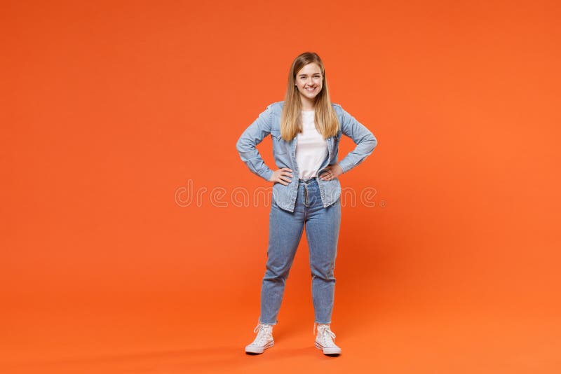 Smiling young woman girl in casual denim clothes posing isolated on orange wall background studio portrait. People emotions lifestyle concept. Mock up copy space. Standing with arms akimbo on waist