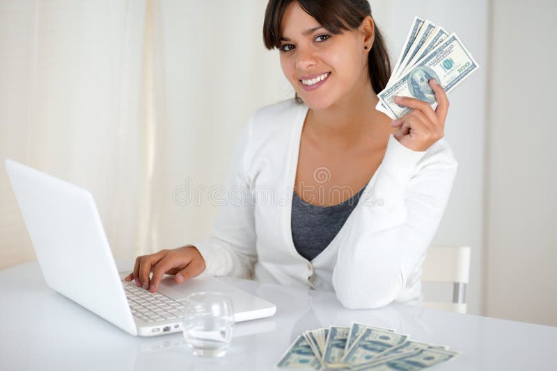 Portrait of a smiling young woman holding up cash money in front of her laptop while is looking at you