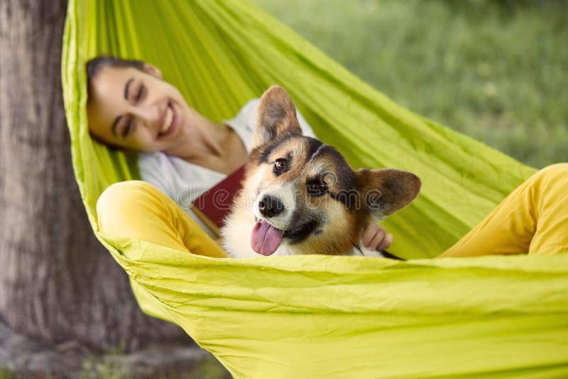 Smiling young woman resting in green hammock with cute dog Welsh Corgi in a park outdoors. Beautiful happy female in white shirt enjoying good day and looking on pet. Concept friendship with dog and human, cute moments, relaxing, carefree. Focus on the dog.