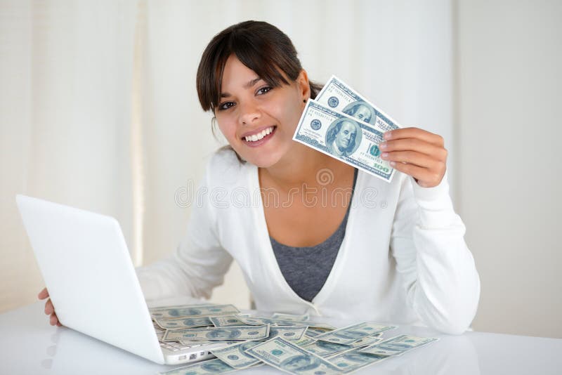 Portrait of a smiling young woman showing you cash money in front of her laptop computer