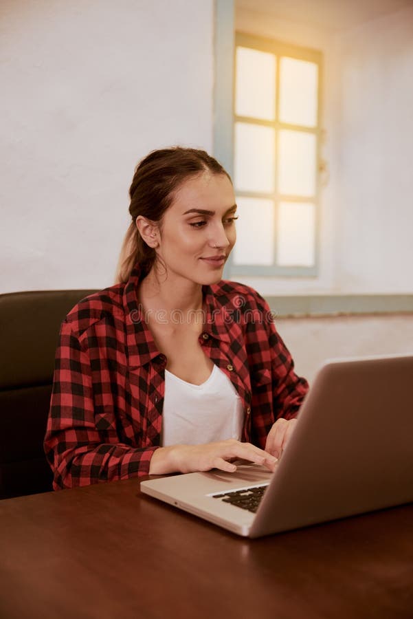 Beautiful young woman typing on her laptop while sitting on an office chair at a desk with a white wall behind her. Beautiful young woman typing on her laptop while sitting on an office chair at a desk with a white wall behind her