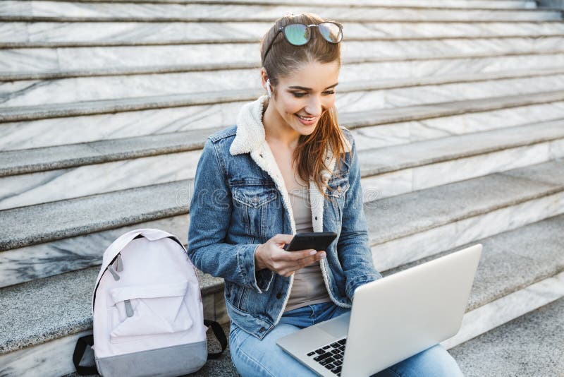 Smiling young woman wearing jacket sitting on stairs outdoors, using laptop computer, holding mobile phone. Smiling young woman wearing jacket sitting on stairs outdoors, using laptop computer, holding mobile phone