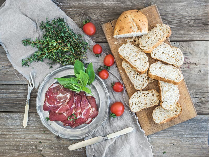 Smoked meat in vintage silver plate with fresh basil, cherry-tomatoes and bread slices over rustic wood