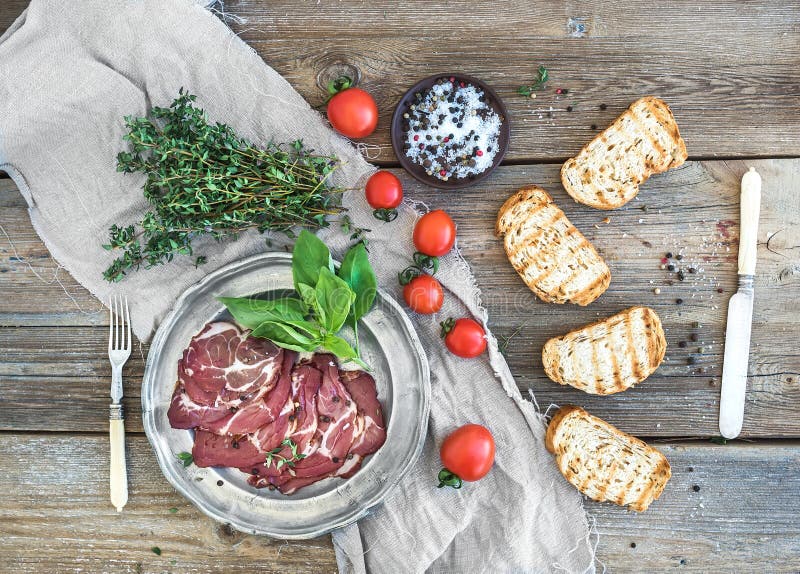 Smoked meat in vintage silver plate with fresh basil, cherry-tomatoes and bread slices over rustic wood