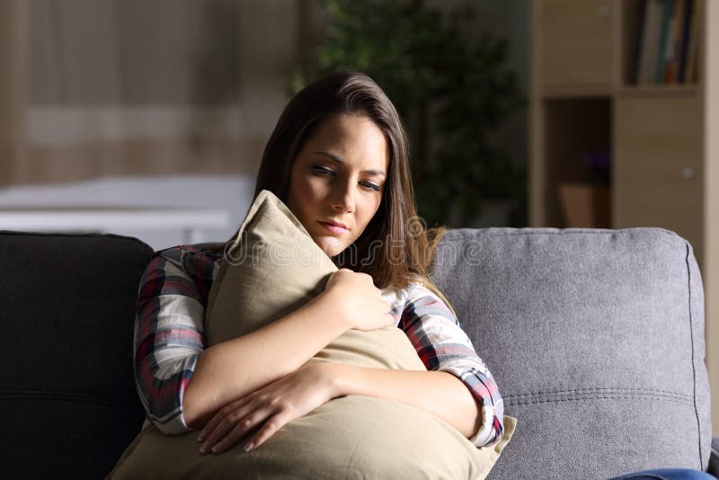Front view of a sad single girl embracing a pillow sitting on a couch in the living room at home with a dark light. Front view of a sad single girl embracing a pillow sitting on a couch in the living room at home with a dark light