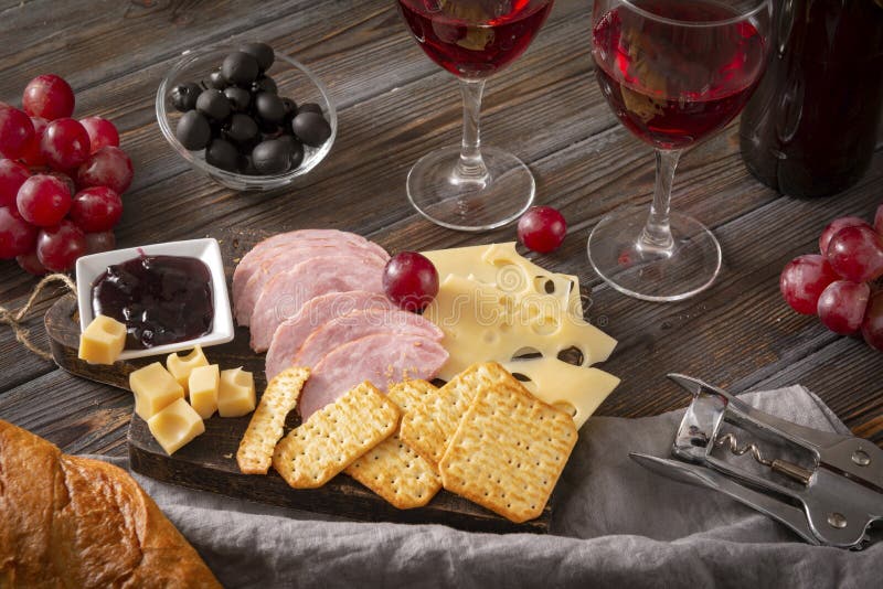 Snack , 2 red wine glasses, slices of cheese , cracker, ham, olives, jam, grapes,  corkscrewon a dark wooden