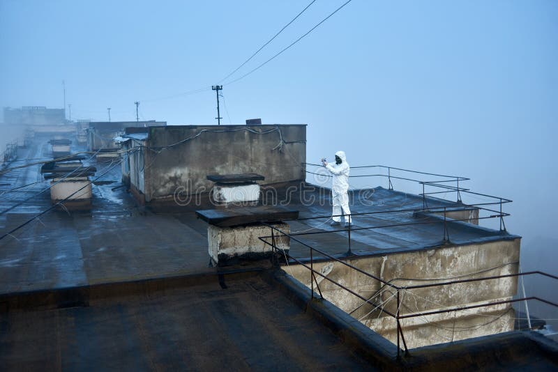 Worker scientist wearing protective coverall and gas mask doing ecological tests on the roof. Snapshot of big flat roof of a block of flats in city, foggy day royalty free stock photography Worker scientist wearing protective coverall and gas mask doing ecological tests on the roof. Snapshot of big flat roof of a block of flats in city, foggy day royalty free stock photography