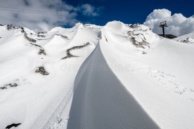 Bank of snow on top of Mount Etna volcano in winter, Sicily island, Italy. Closeup of snowdrift on volcano slopes with black lava stones under blue sky, clouds and smoke. Cable car road in background. Bank of snow on top of Mount Etna volcano in winter, Sicily island, Italy. Closeup of snowdrift on volcano slopes with black lava stones under blue sky, clouds and smoke. Cable car road in background.