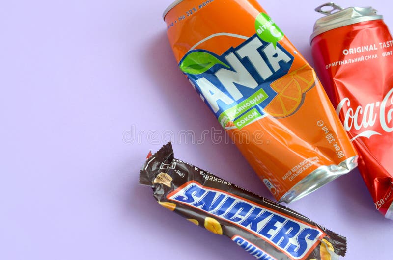 Snickers chocolate bar wrapping lies with empty red Coca Cola and orange Fanta tin cans on lilac pastel background. Famous drink