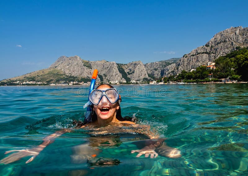 A view of a funny face - young girl with diving sea mask on holiday diving - snorkeling blue - green Adriatic sea - Croatia-Dalmatia. In background is mountain cliff and town Omis Horizontal color photo. A view of a funny face - young girl with diving sea mask on holiday diving - snorkeling blue - green Adriatic sea - Croatia-Dalmatia. In background is mountain cliff and town Omis Horizontal color photo.