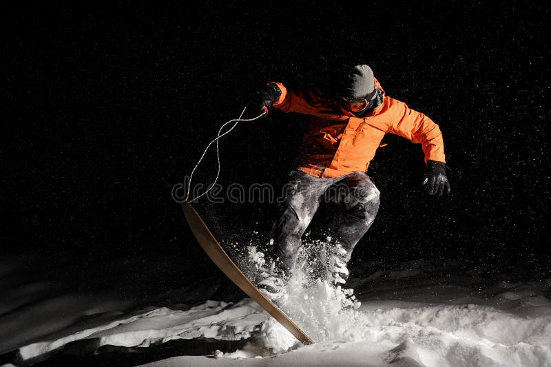 Snowboarder maschio professionista in abiti sportivi arancio che saltano sulla neve alla notte immagine stock Snowboarder maschio professionista in abiti sportivi arancio che saltano sulla neve alla notte immagine stock