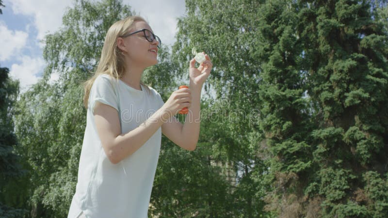 Snygg ung kvinna i glasögon som blåser soapbubblor som flyger bort i parken framför träd på vardagssommaren