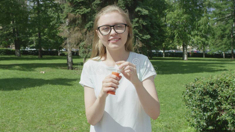Snätt blond flicka i glas som blåser små tvålbubblor i parken och leker med glädje vid kameran på solig sommardag.
