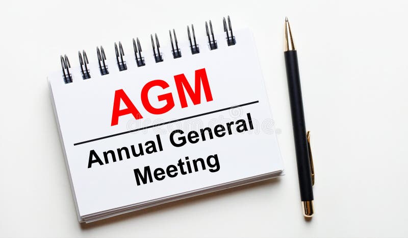 On a light background, a white notebook with are words AGM Annual General Meeting and a pen. On a light background, a white notebook with are words AGM Annual General Meeting and a pen.