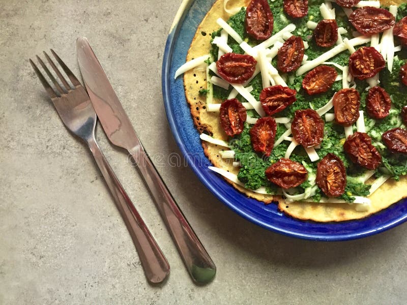 Top-down view of homemade socca on a blue plate topped with homemade green pesto (actually made with nettles but could be any green such as spinach or kale), slow-roasted grape tomatoes and shredded white mozzarella cheese. Fork and knife next to plate. Socca, also known as farinata, is a gluten-free crepe or flatbread made with chickpea flour that's popular in southern France. Vegetarian meal. Top-down view of homemade socca on a blue plate topped with homemade green pesto (actually made with nettles but could be any green such as spinach or kale), slow-roasted grape tomatoes and shredded white mozzarella cheese. Fork and knife next to plate. Socca, also known as farinata, is a gluten-free crepe or flatbread made with chickpea flour that's popular in southern France. Vegetarian meal.