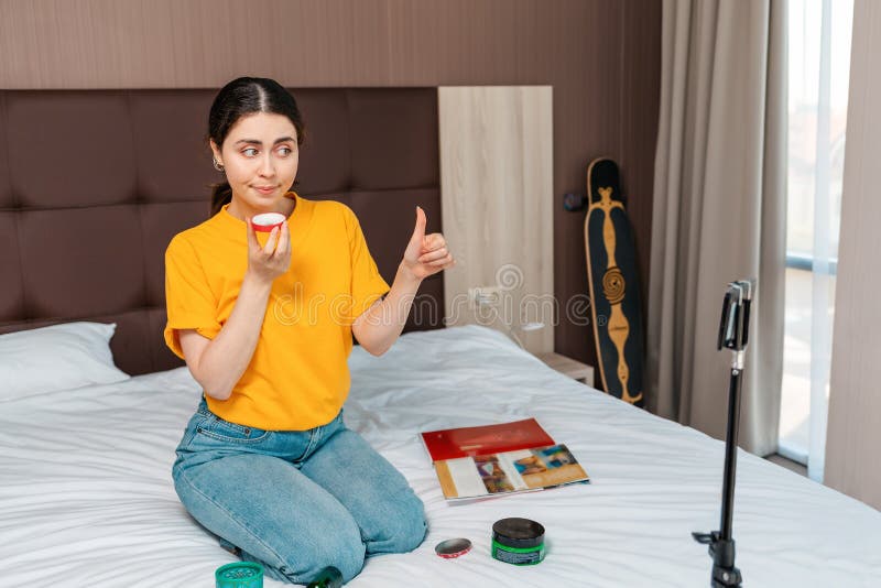 Social media. A young female vlogger streaming a live video to her smartphone sitting on bed and thumbs up showing cosmetics. Home related. Social media. A young female vlogger streaming a live video to her smartphone sitting on bed and thumbs up showing cosmetics. Home related.