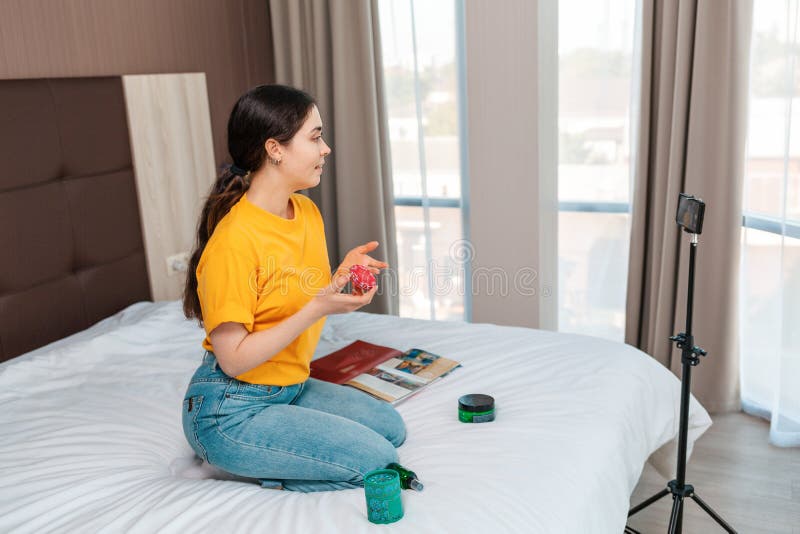 Social media. A beautiful female vlogger streaming a live video to her smartphone sitting on bed and talking about cosmetics. Side view. Windows on the background. Social media. A beautiful female vlogger streaming a live video to her smartphone sitting on bed and talking about cosmetics. Side view. Windows on the background.