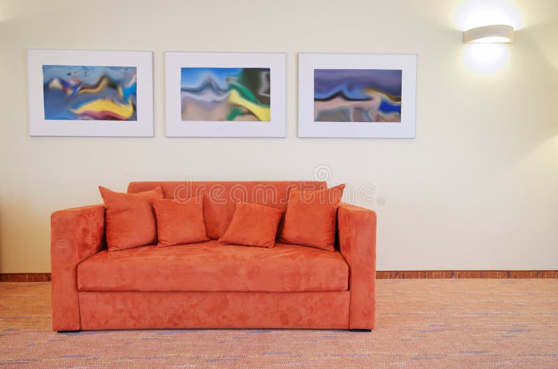 An orange sofa standing alone at the wall of a hotel room, three pictures on the wall, carpeted floor. An orange sofa standing alone at the wall of a hotel room, three pictures on the wall, carpeted floor.