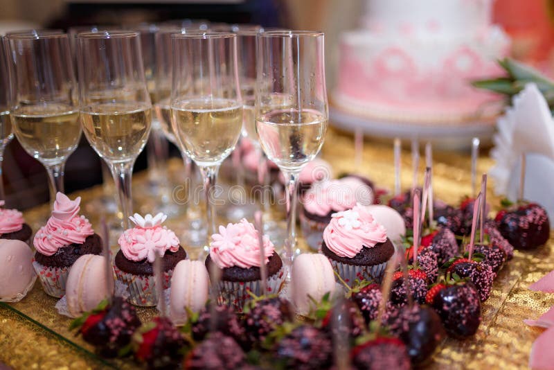 festive candy bar with cupcakes, champagne glasses and strawberries in chocolate glaze. Unfocused pink cake on background. Weddin