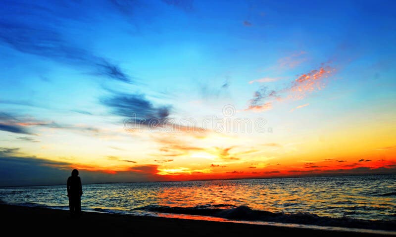 Ein stimmungsvolles und spektakuläres australischen tropischen Sonnenuntergang über dem Meer, mit einer schwarzen silhouette Bild von einer Frau Stand am Rand des Wassers allein, mit Blick auf all die Natürliche Schönheit des Meeres und die herrliche, lebendige array von Farben.