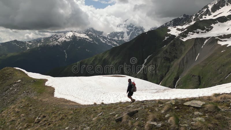 Solo hiker with backpack walks on snowy mountain trail. Man trekking in high peaks, exploring nature. Aerial view of stock video